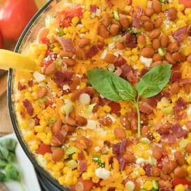 Jiffy cornbread salad layered with corn, beans, cheese, bacon, green onions, and fresh basil leaves in a clear glass bowl.