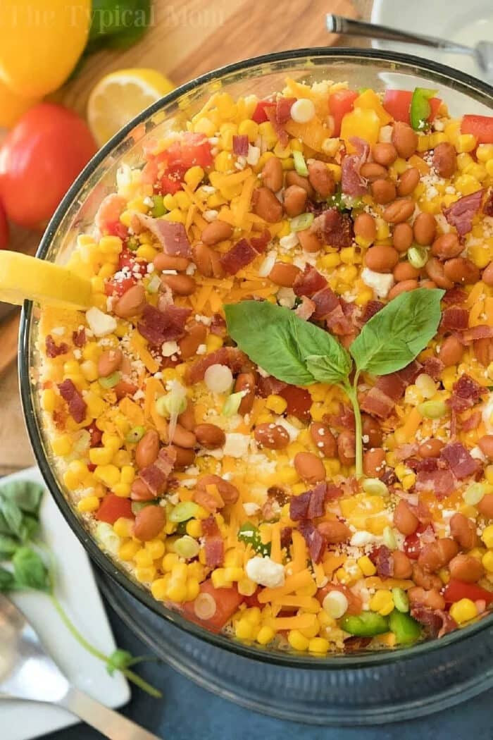 Layered cornbread salad is a perfect side dish during the holidays or a barbecue! Layers of corn, cheese and more make it irresistible! #corn #salad #layered #cornbread #sidedish Jiffy cornbread salad layered with corn, beans, cheese, bacon, green onions, and fresh basil leaves in a clear glass bowl.