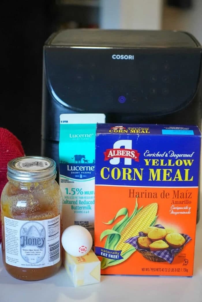 A carton of buttermilk, cornmeal box, honey jar, egg, and butter stick are arranged in front of an air fryer—perfect ingredients for making delicious air fryer cornbread.