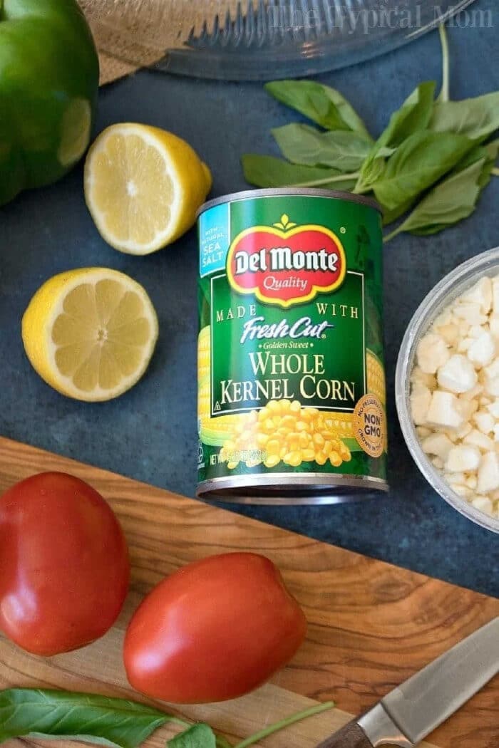 This easy layered cornbread salad recipe is a perfect side dish during the holidays or a barbecue! Layers of corn, cheese and more make it irresistible! #corn #salad #layered #cornbread #sidedish A can of Del Monte whole kernel corn surrounded by tomatoes, lemon, basil, and chopped onions—perfect ingredients for a fresh Jiffy cornbread salad.