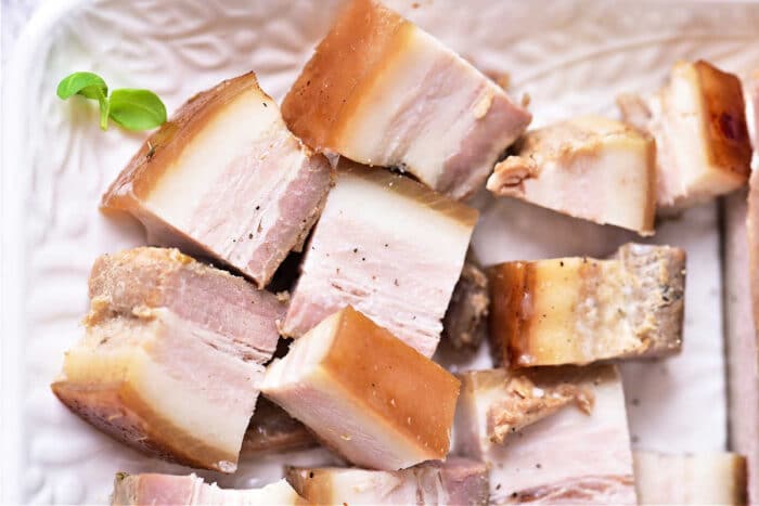 Cubes of instant pot pork belly with visible fat layers on a white textured plate, garnished with a green herb.