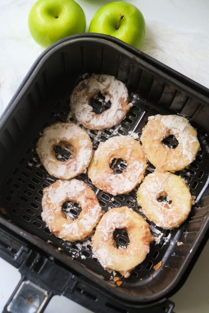 Sliced air fryer apples coated in flour are arranged in the basket, with two fresh green apples nearby.