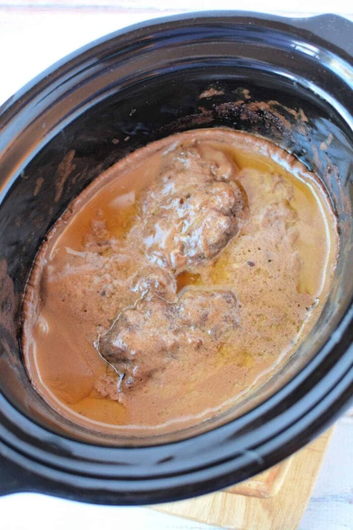 Two pieces of Crockpot Salisbury Steak simmer in rich brown gravy inside a black slow cooker, filling your kitchen with mouthwatering aromas.