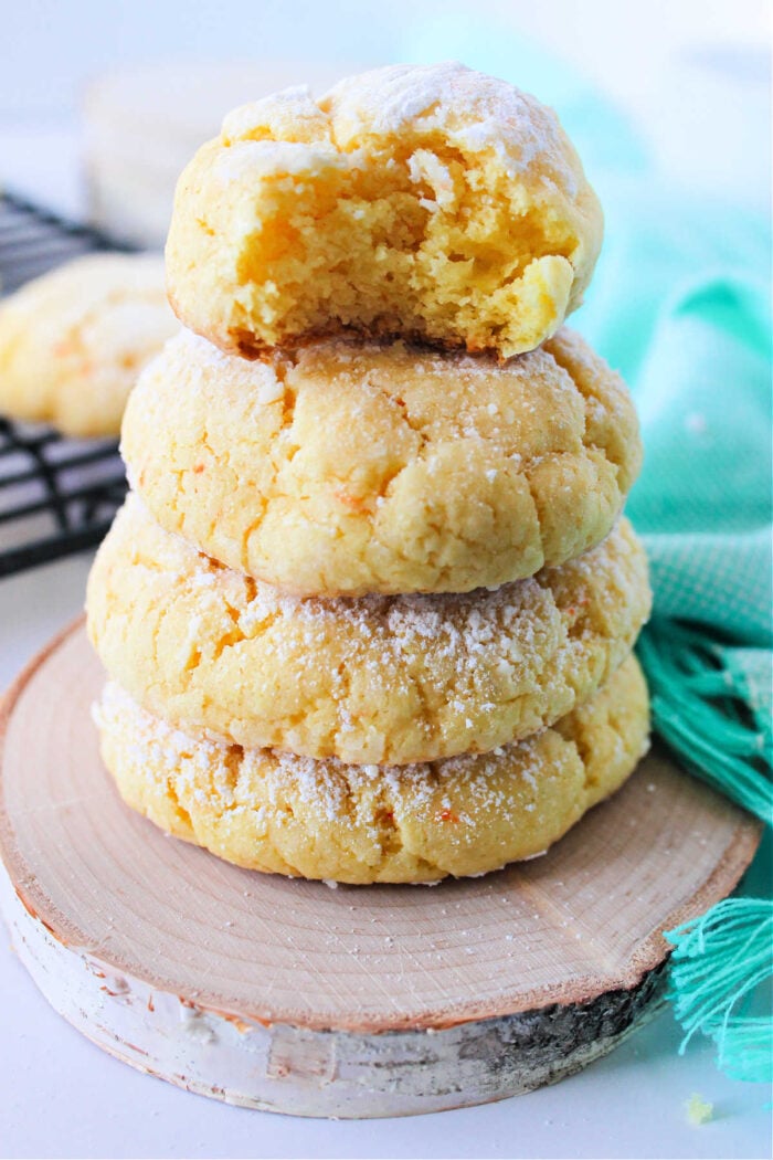 A stack of four Cake Mix Butter Cookies, dusted with powdered sugar, with the top cookie bitten, sits on a wooden coaster.