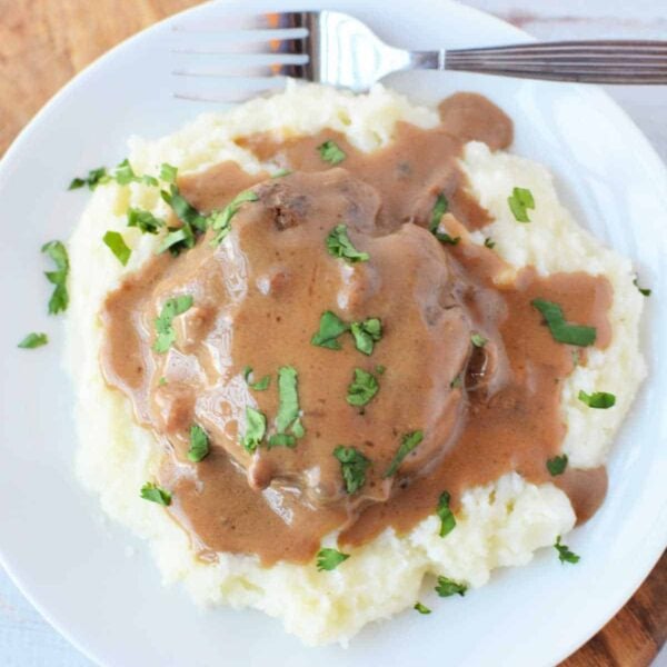Mashed potatoes topped with brown gravy and chopped parsley, served alongside Crockpot Salisbury Steak on a white plate with a fork.