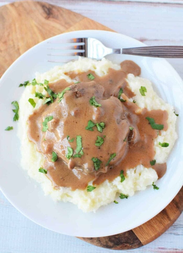 Mashed potatoes topped with brown gravy and chopped parsley, served alongside Crockpot Salisbury Steak on a white plate with a fork.