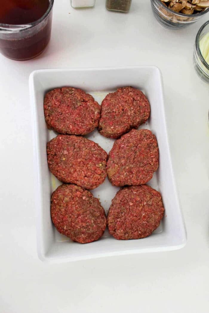 Six raw hamburger steak patties arranged in a white rectangular dish on a white surface.