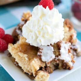 A slice of apple spice dump cake topped with whipped cream and a raspberry on a white plate, with more raspberries on the side.