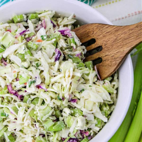 Bowl of Blue Cheese Coleslaw with a wooden fork, surrounded by lemon halves and celery stalks.