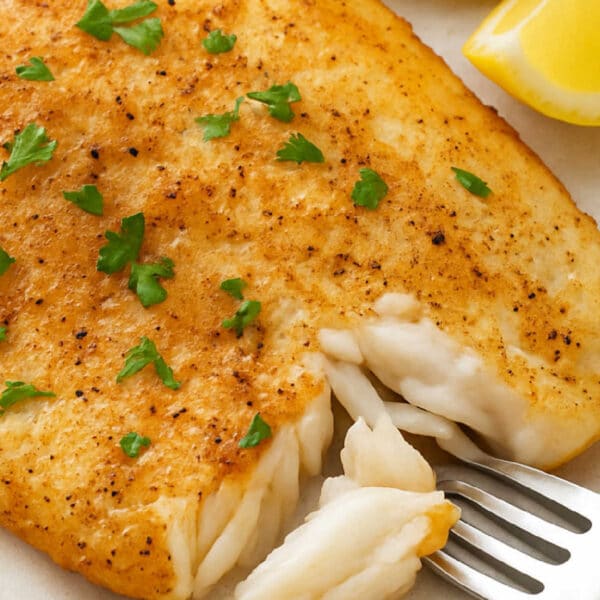 Golden-brown baked fish fillet, cooked from frozen tilapia in the air fryer, with parsley, lemon wedges, and a fork breaking off a piece.
