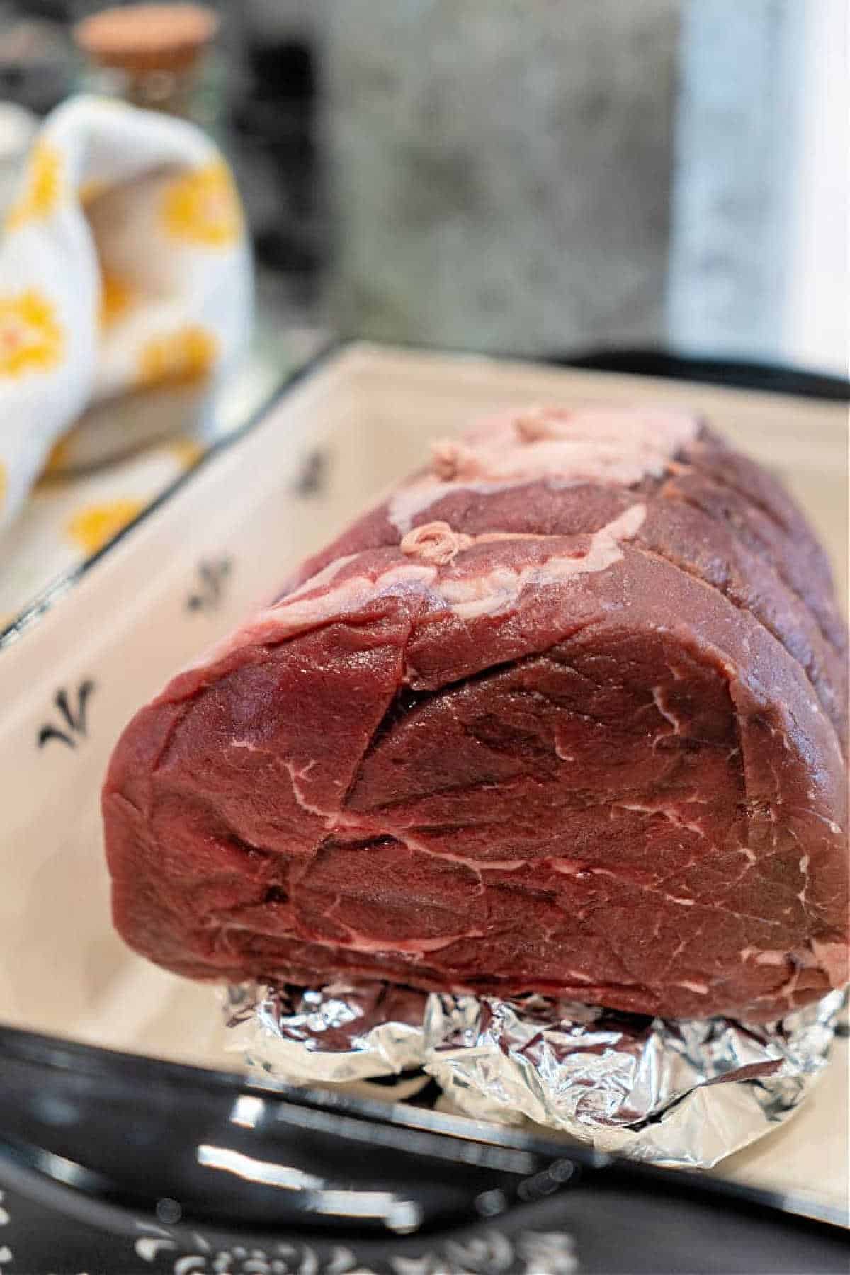 A raw top round roast sits on foil in a ceramic baking dish, ready to be cooked&mdash;perfect for your next Top Round Roast Recipe.