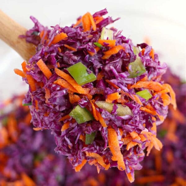 Close-up of a wooden spoon holding shredded red cabbage coleslaw with carrots and green bell pepper.