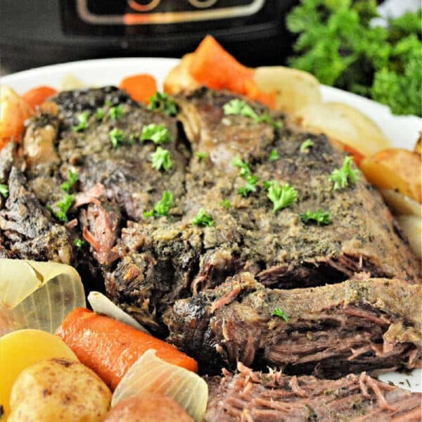 A plate of pot roast with potatoes, carrots, and onions, garnished with parsley, sits in front of a slow cooker—perfect for making a frozen roast in slow cooker for a hearty meal.