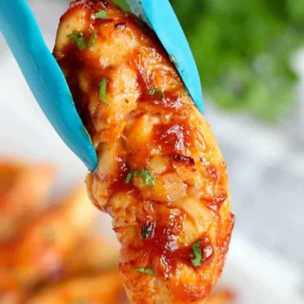 A piece of saucy air fryer chicken tenders no breading held by blue tongs, with herbs sprinkled on top.