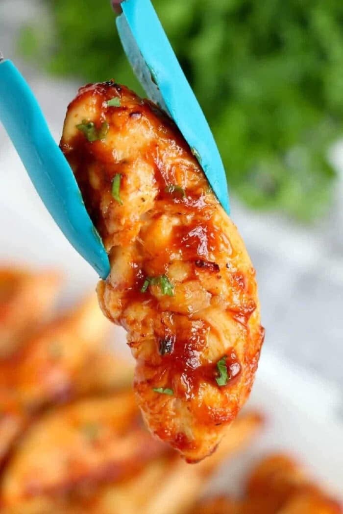 A piece of saucy air fryer chicken tenders no breading held by blue tongs, with herbs sprinkled on top.