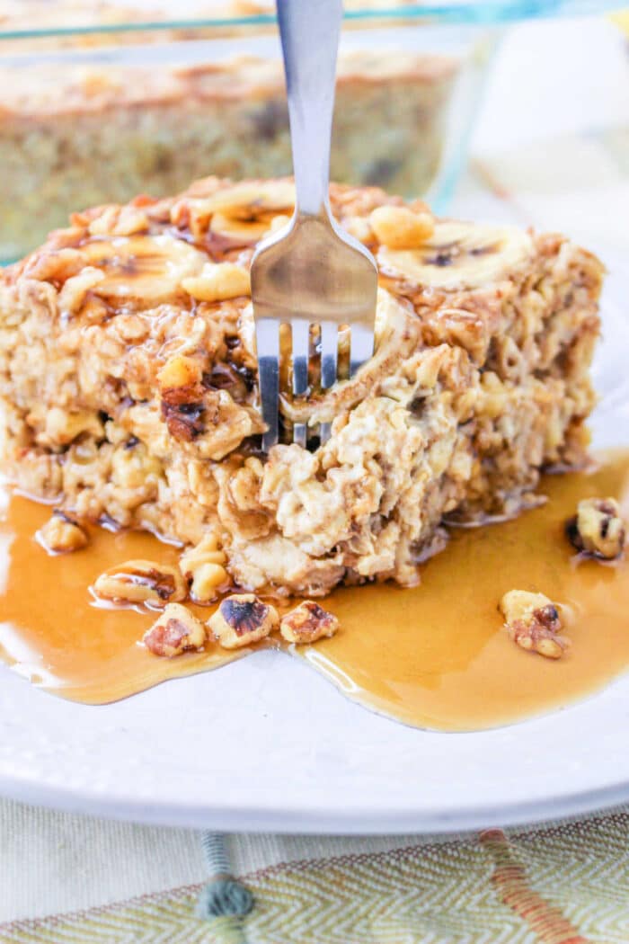 A fork digging into a slice of Bananas and Cream Oatmeal, baked to perfection and topped with syrup and chopped nuts on a white plate.