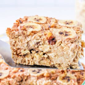 A spatula lifts a square of Banana baked Oatmeal, topped with fresh banana slices and crunchy nuts.