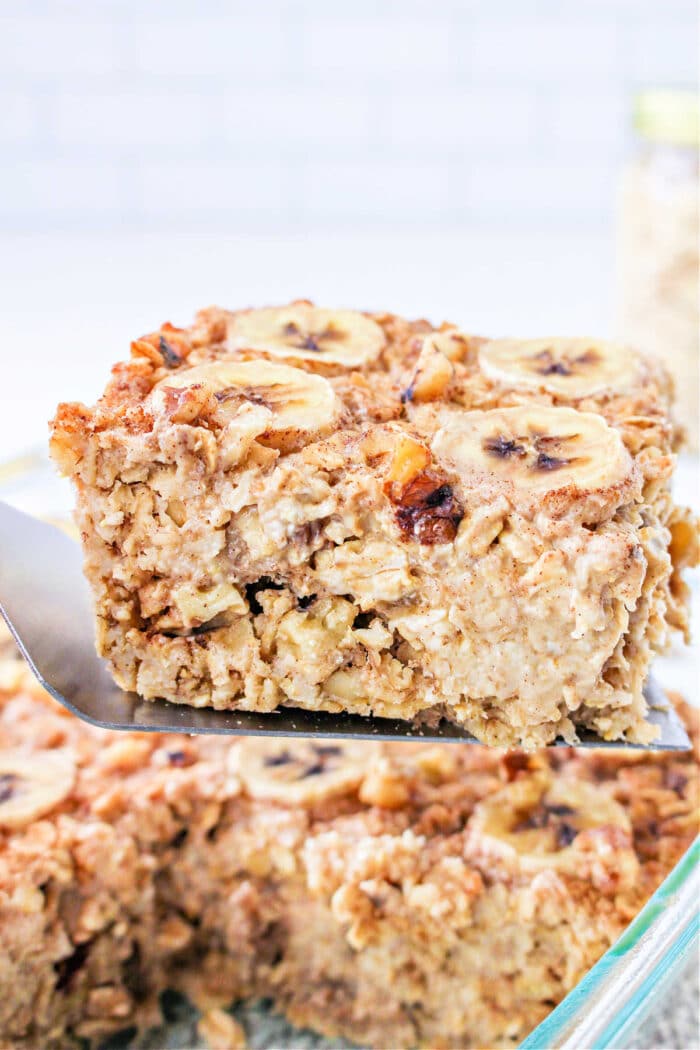 A spatula lifts a square of Banana baked Oatmeal, topped with fresh banana slices and crunchy nuts.