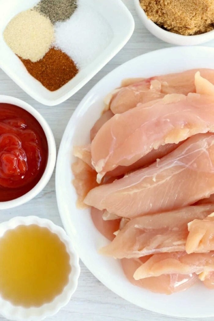 Raw chicken strips on a plate, ready to become delicious air fryer chicken tenders with no breading, are surrounded by bowls of ketchup, spices, brown sugar, and a yellow liquid for flavor.