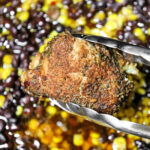 Metal tongs hold a seasoned piece of crockpot jerk chicken above a mix of black beans and corn.