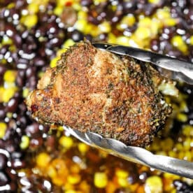 Metal tongs hold a seasoned piece of crockpot jerk chicken above a mix of black beans and corn.