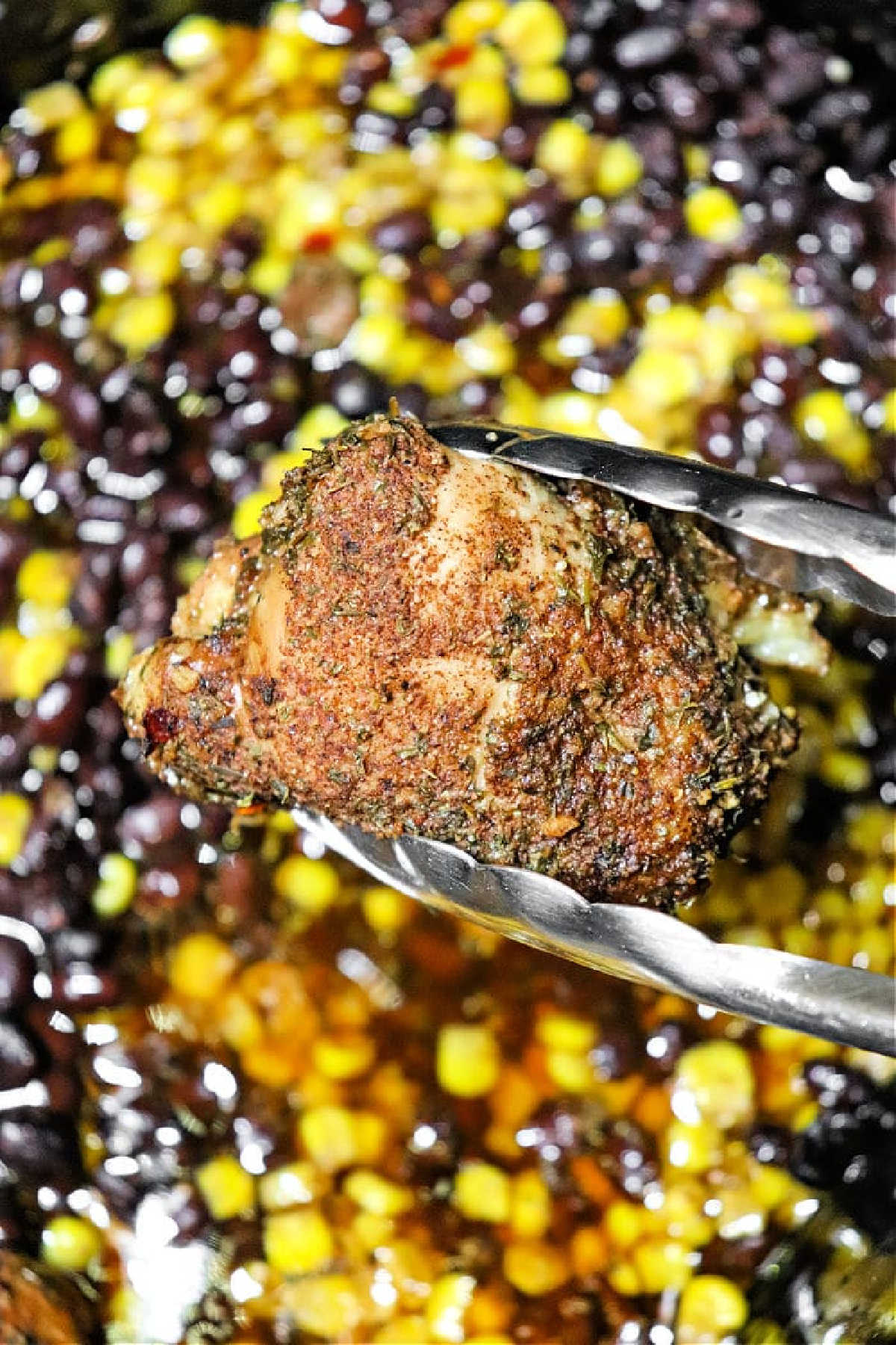 Metal tongs hold a seasoned piece of crockpot jerk chicken above a mix of black beans and corn.