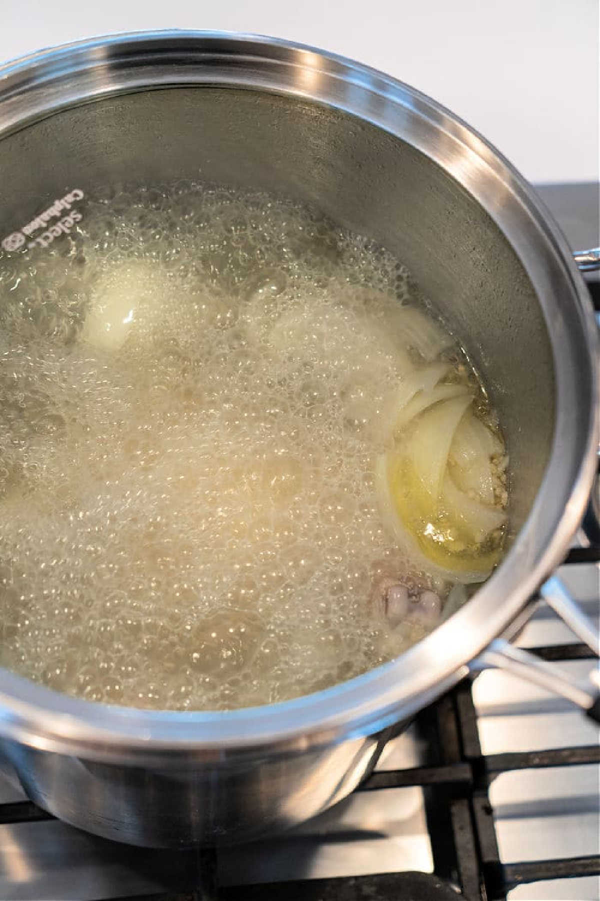 A pot of boiling water with onion slices and other ingredients simmers on the stovetop, starting the base for a flavorful whole chicken soup.
