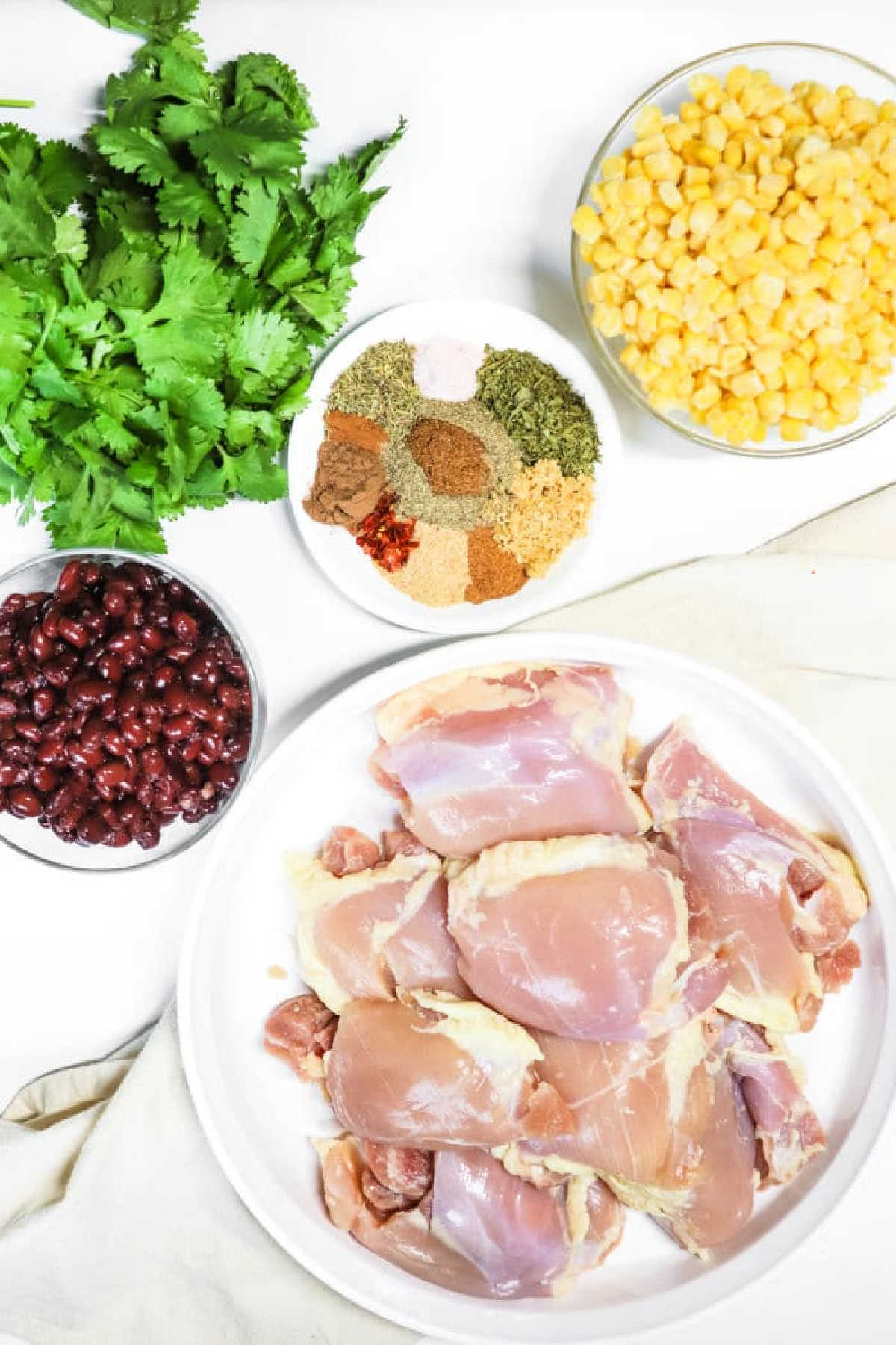 Raw chicken thighs, black beans, corn, cilantro, and assorted spices arranged in bowls on a white surface—perfect for making flavorful crockpot jerk chicken.