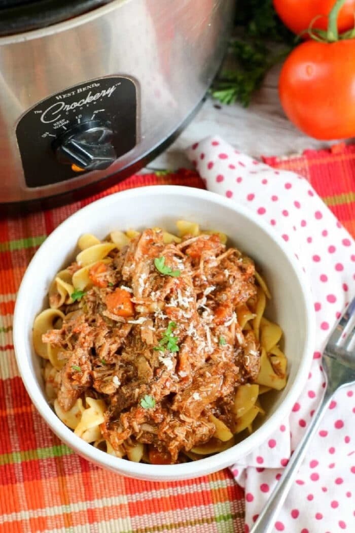 Bowl of pasta topped with shredded beef ragu made from slow cooker spaghetti sauce with leftover beef, beside fresh tomatoes on a colorful placemat.