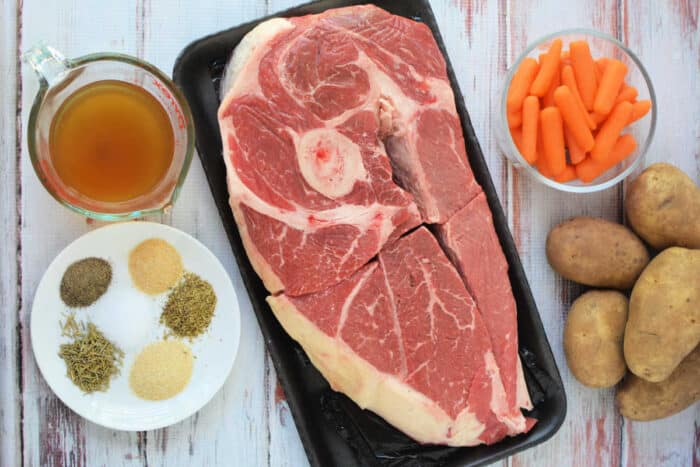 Raw chuck roast, baby carrots, potatoes, broth, and assorted spices—perfect ingredients for a comforting slow cooker meal—are arranged on a white wooden surface.