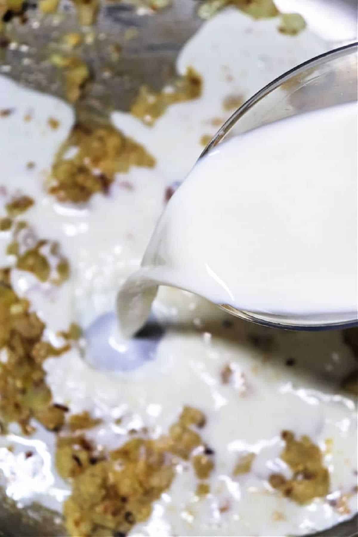 Milk is poured from a glass jug into a pan with sautéed onions and spices, showing an essential step in how to make gravy from chicken drippings for a rich, creamy finish.