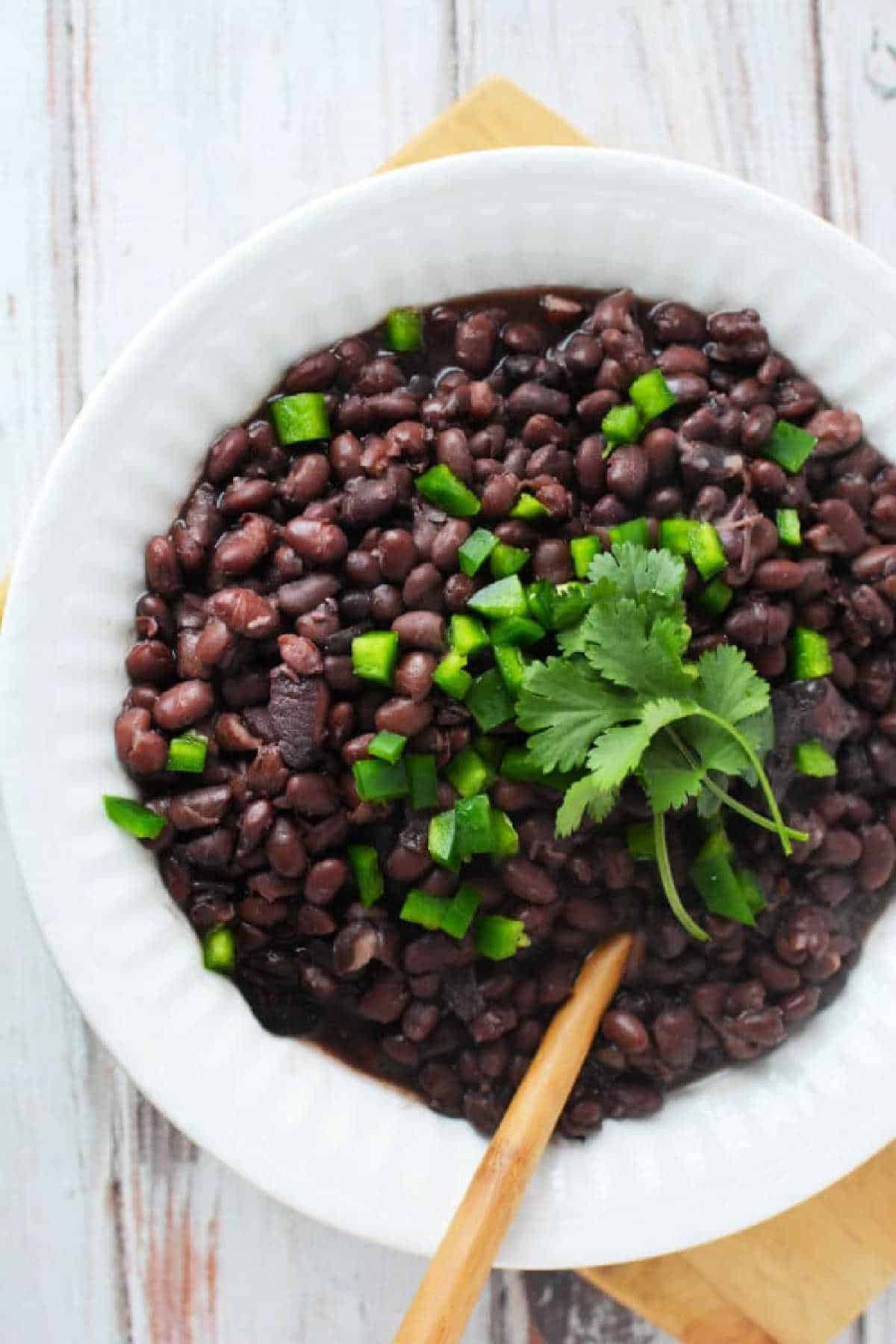 Slow Cooker Black Beans in a white bowl, garnished with chopped green peppers and cilantro, served with a wooden spoon.
