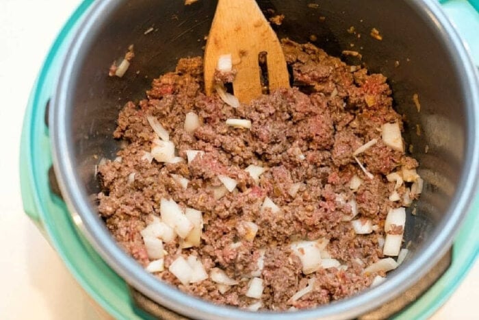 Ground beef and chopped onions being cooked in a pot with a wooden spatula for a delicious pressure cooker lasagna casserole.