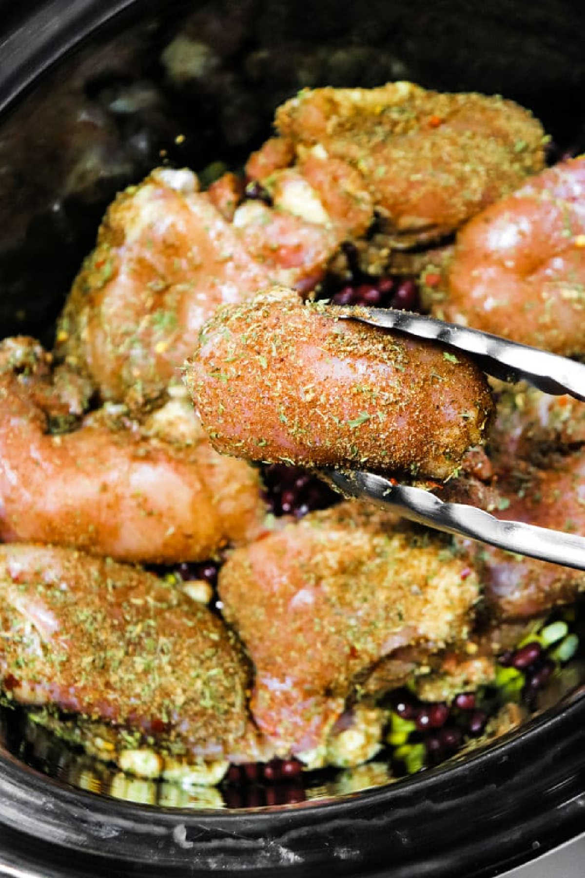 Tongs holding seasoned raw chicken above more chicken in a crockpot jerk chicken dish with beans and vegetables.