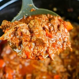 A ladle lifting shredded beef in a rich, red slow cooker spaghetti sauce with leftover beef.