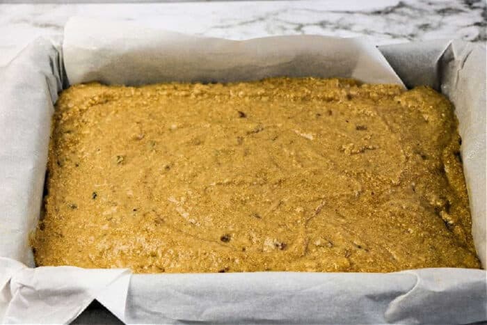 A rectangular baking pan lined with parchment paper is filled with unbaked applesauce spice cake batter, resting on a marble countertop.