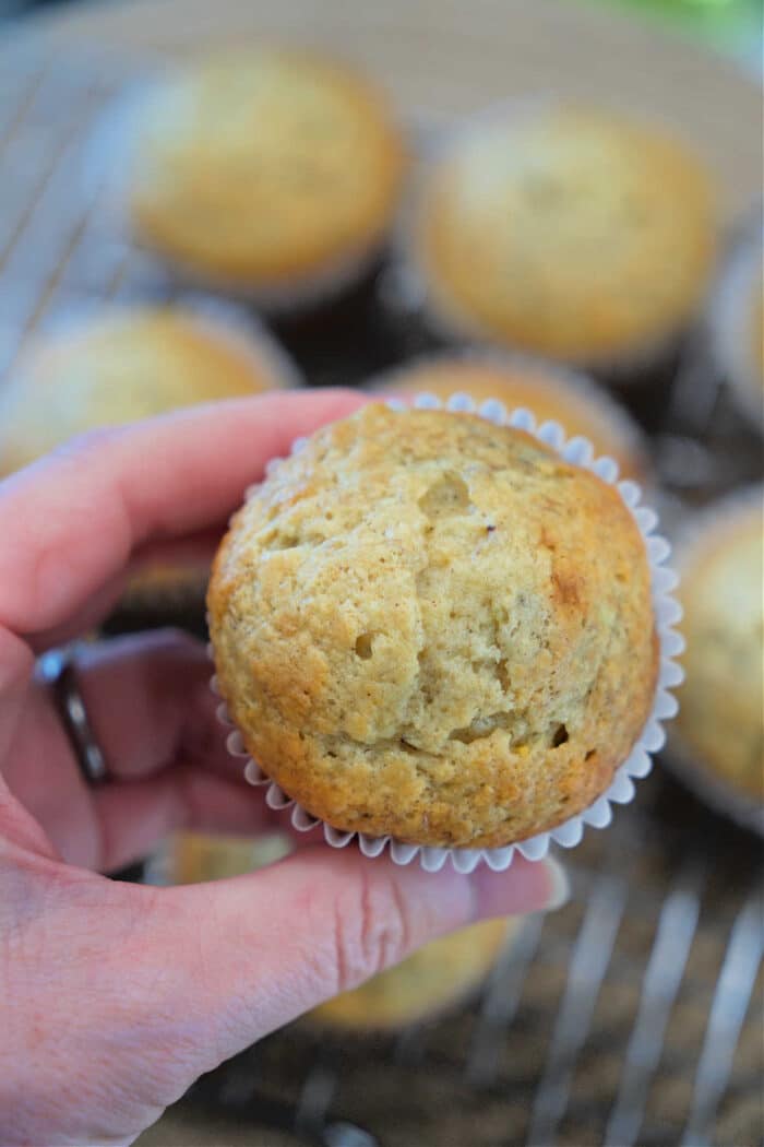 A hand holds a freshly baked banana muffin in a white paper liner, with more banana muffins on a cooling rack behind.