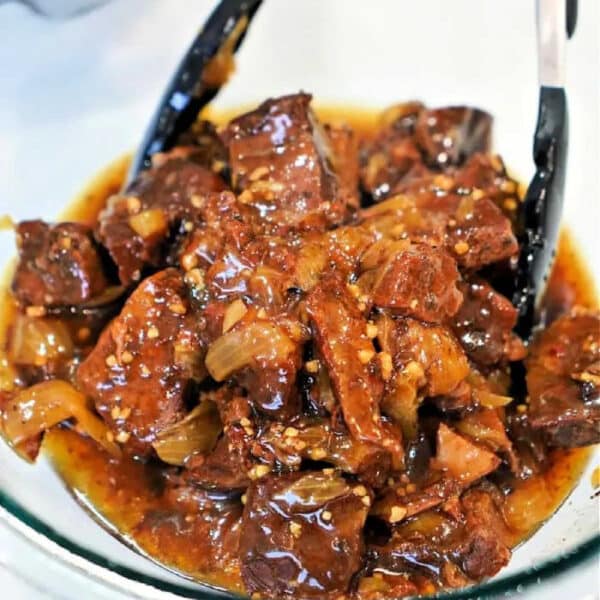 Chunks of braised beef, prepared using a classic beef arm roast recipe, in a rich, glossy sauce with onions, served in a glass bowl with tongs.
