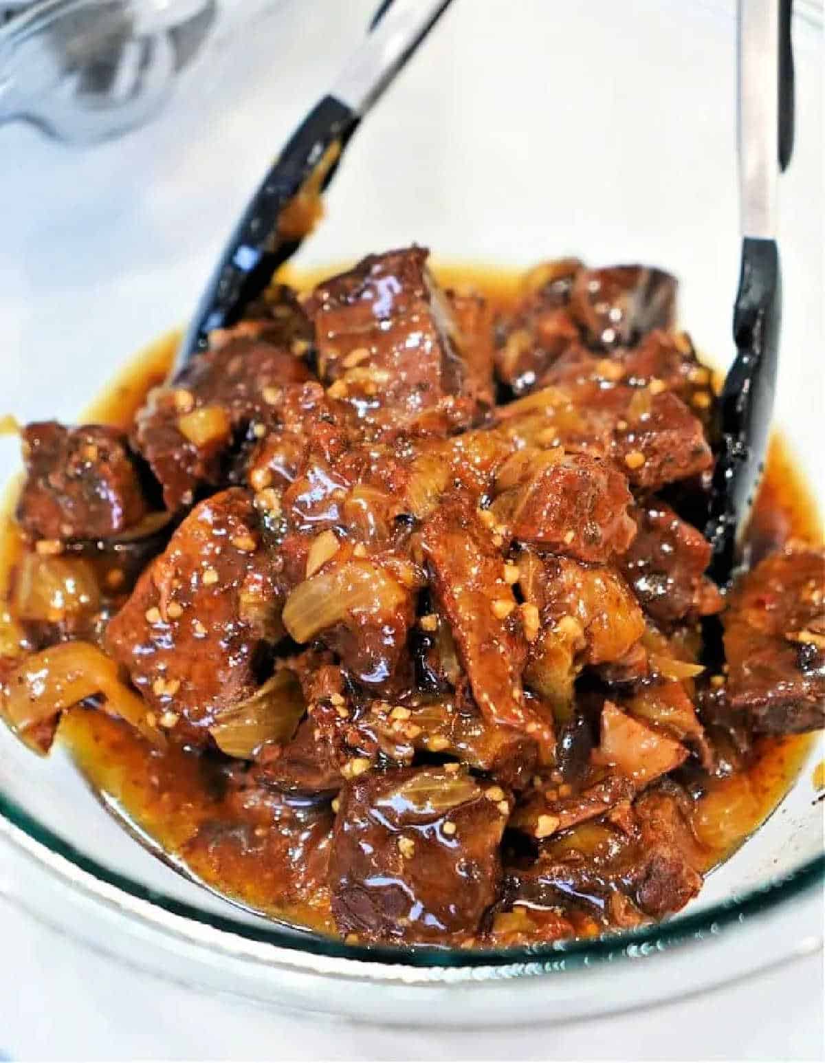 Chunks of braised beef, prepared using a classic beef arm roast recipe, in a rich, glossy sauce with onions, served in a glass bowl with tongs.
