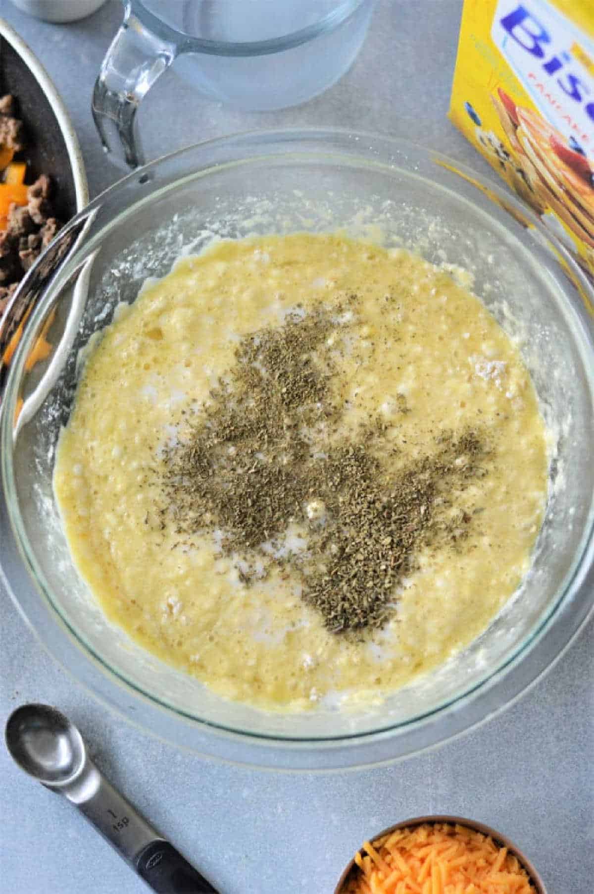 A glass bowl with batter and dried herbs, surrounded by cheese, Bisquick mix, and a measuring spoon—perfect ingredients for making a delicious Breakfast Casserole with Bisquick.