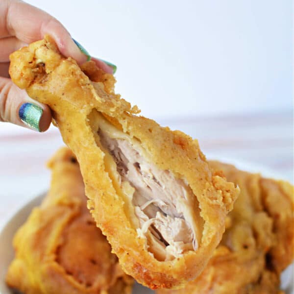 A hand holds a crispy pan fried chicken drumstick with a bite taken out, exposing the juicy inside; plate in background.