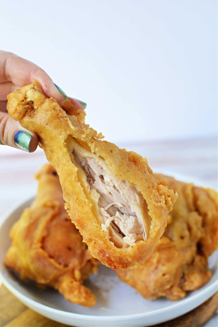 A hand holds a crispy pan fried chicken drumstick with a bite taken out, exposing the juicy inside; plate in background.