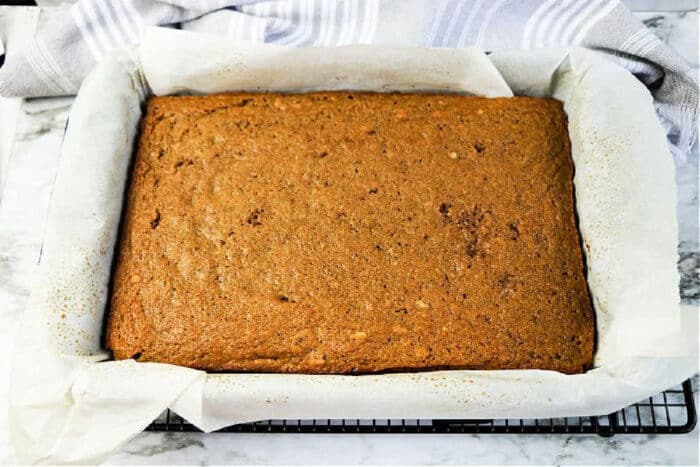 A rectangular applesauce spice cake rests in a parchment-lined baking pan on a cooling rack.