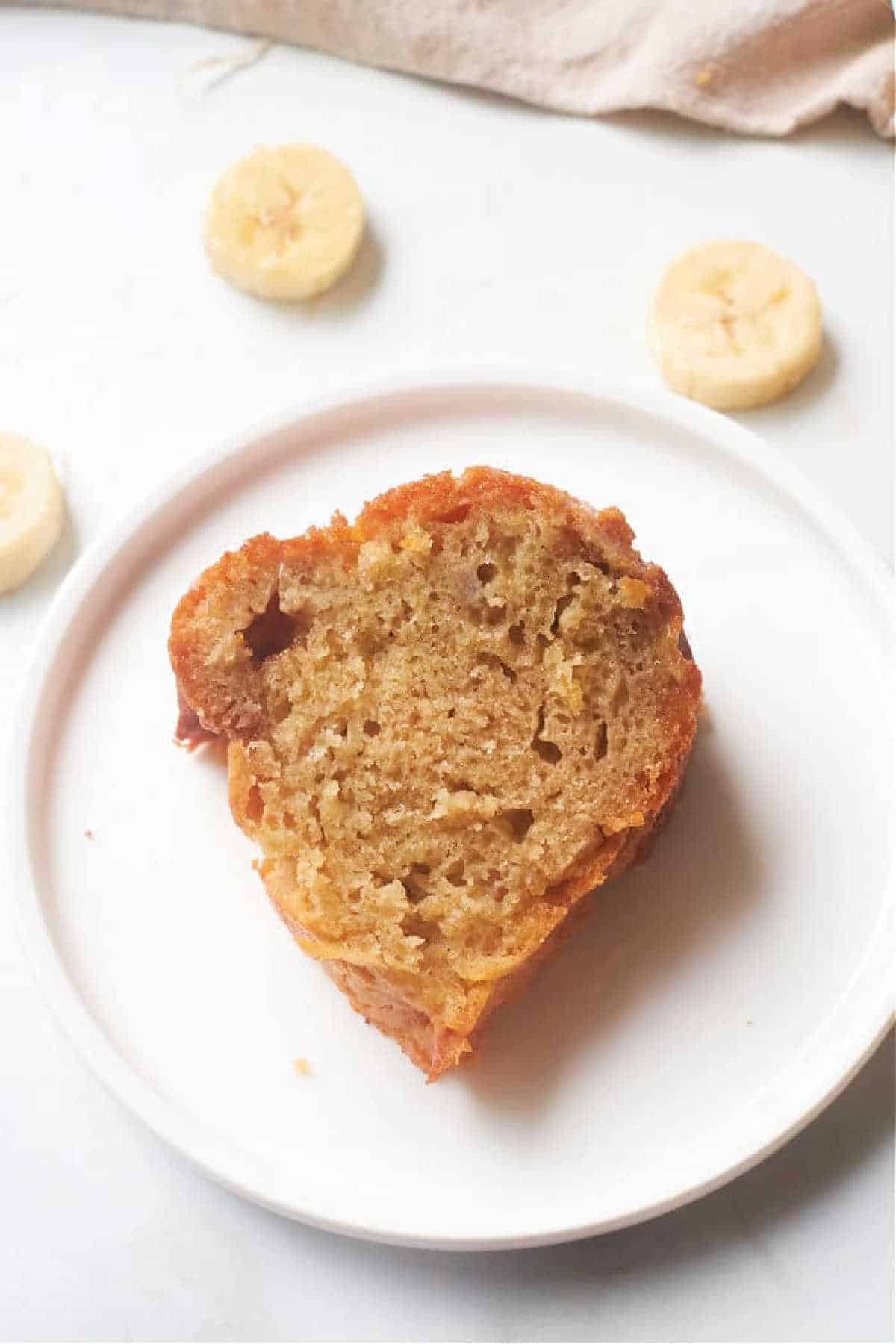 A slice of Banana Bread Bundt Cake sits on a white plate with banana slices in the background.