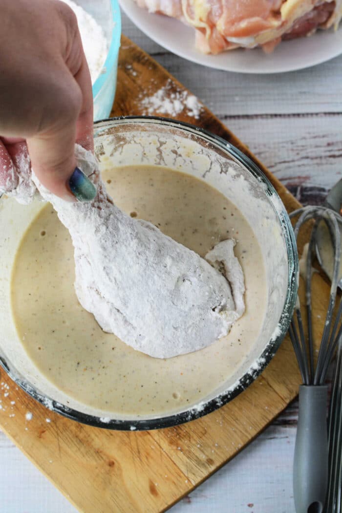 A hand dips a floured chicken drumstick into a bowl of batter on a wooden cutting board, preparing it for delicious pan fried chicken.