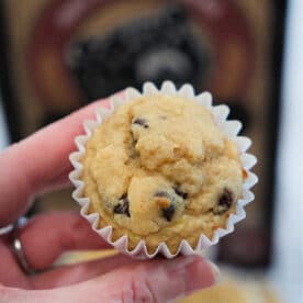 A hand holding a Kodiak banana chocolate chip muffin in a white paper liner, with more banana muffins and a box in the background.