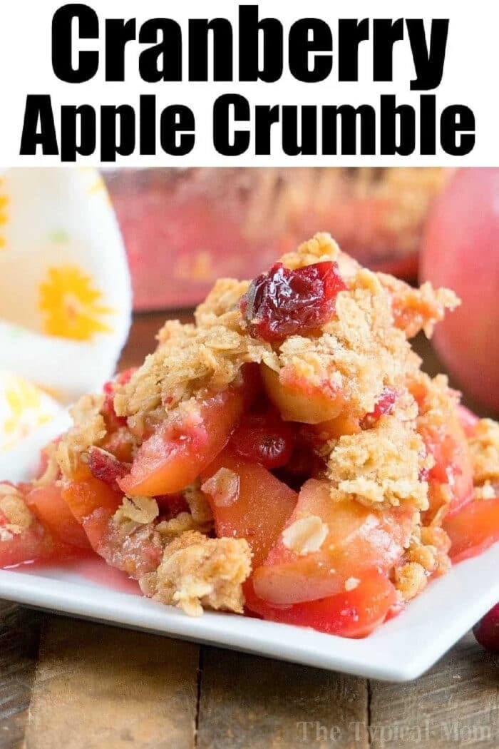 A square plate with cranberry apple cobbler crumble topped with oats, set on a wooden table with fruit in the background.