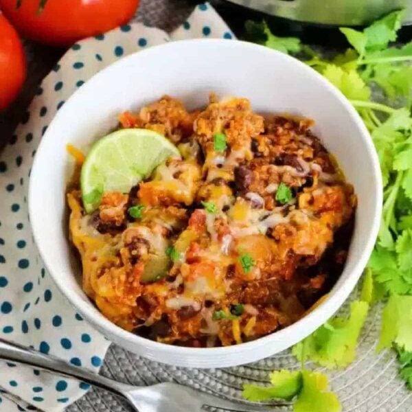A bowl of cheesy chili with beans, topped with a lime wedge, sits beside tomatoes and fresh cilantro—perfect comfort food reminiscent of a crockpot enchilada casserole.
