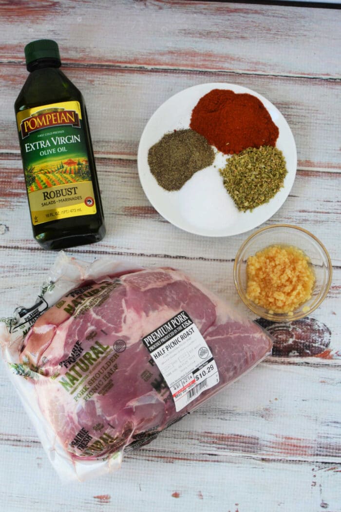 Pork picnic Roast Recipe, with a bottle of olive oil, minced garlic, and a plate of herbs and spices on a white wood surface—perfect ingredients before roasting in the oven.