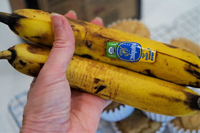 A hand holds two ripe bananas with stickers, in front of a wire rack filled with freshly baked Kodiak Banana Muffins.